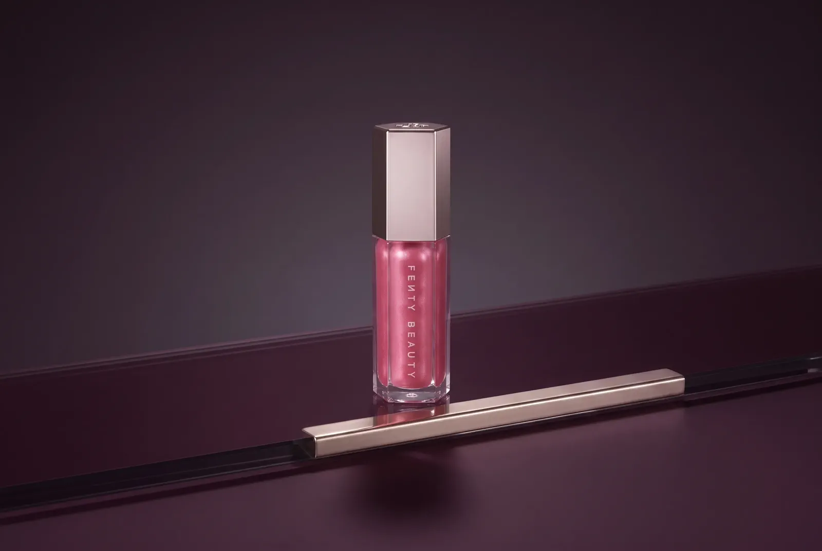 A faceted pink lip-gloss tube in a polished plum studio with controlled reflective highlights.